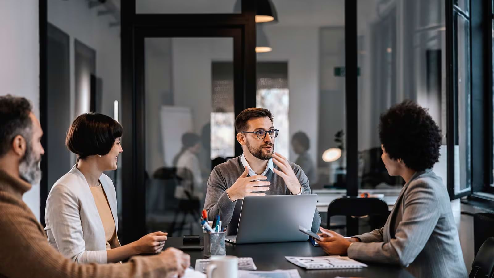 A group of diverse professionals engaged in a collaborative discussion around a table, with laptops and notebooks visible, in a modern office setting. The glass walls provide a view of the bustling workplace environment beyond, showcasing an atmosphere of teamwork and productivity.